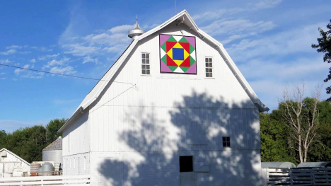 A white barn with a colorful barn quilt at the peak on the front side.