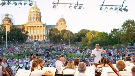 With the Iowa State Capitol in the background, a crowd of people sit on the Capitol lawn looking on at the Des Moines Symphony playing. The Symphony members are in the foreground, facing away from the viewer and toward the conductor who is leading the symphony.