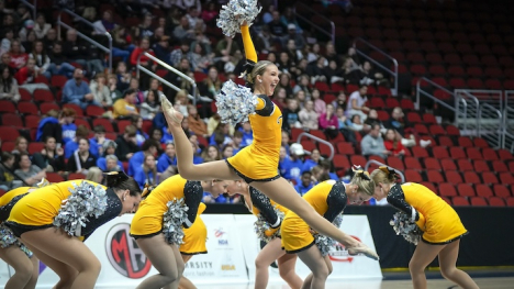 Female dancers performing in yellow uniforms with silver pom poms. The dancer in the center foreground is leaping in the air with her arms raised.