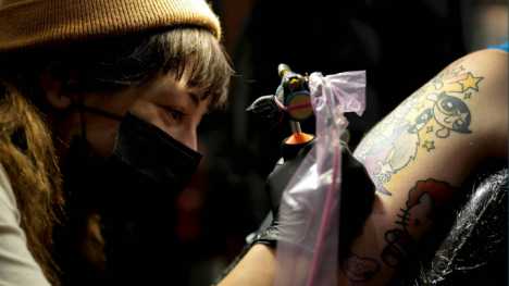 A masked tattoo artist adding a tattoo to a client's arm
