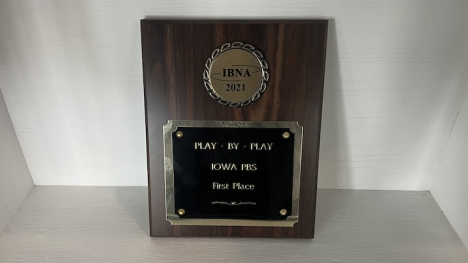 An image of a 2021 IBNA Award, a wooden plaque with bronze and black metal. At the top is a circular bronze piece with the inscription "IBNA 2021." Below is a black plate surrounded by bronze with the inscription "Play-by-Play, Iowa PBS, First Place,"