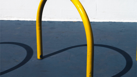 A curved, yellow bike rack casting a shadow on the concrete ground.
