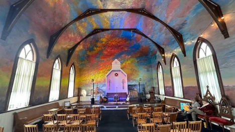 Interior of a chapel with a colorful painted ceiling, arched windows, wooden chairs, and a small stage with a church facade backdrop.