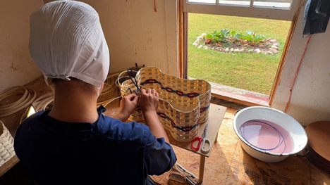 A young Amish woman with her back to the camera making a woven basket.