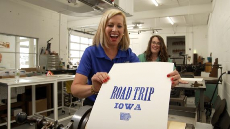 Host Brooke Kohlsdorf holding a "ROAD TRIP IOWA" poster