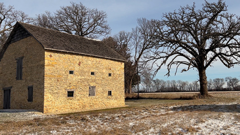 A tall, two-story, rectangular, stone building with a shingled roof stands on snowy ground in rural Iowa.