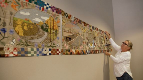 An individual in a white shirt hanging a colorful, quilted fabric art piece on a wall. 
