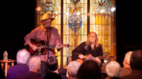 Two musicians with guitars perform in front of a stained glass window.