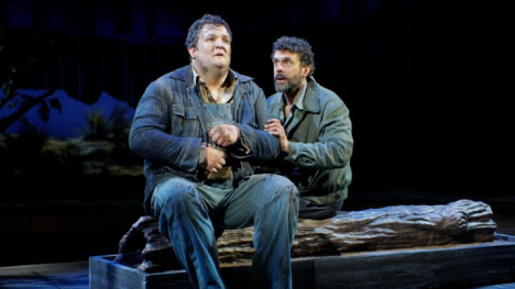 Two actors sit on a wooden log, one appearing distressed in denim overalls, while the other, in a gray jacket, leans in with a concerned expression.