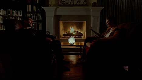 A man and woman sitting in chars opposite each other in front of a fireplace in a dimly lit room.