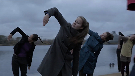 Dancers in motion (leaning left with right arm up, bent at the elbow and hand pointing down) outside by water.