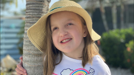 A young girl with blond hair and a sun hat on stands next to a palm tree.