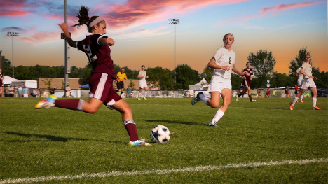 A girls high school soccer game - a player in maroon runs up to the soccer ball to kick it downfield.