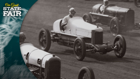 Racers in open-top cars wearing goggles race on a dirt track