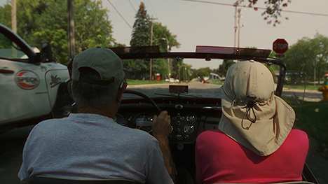 An older man and woman are seen driving down the road in a convertible ... as viewed from the backseat.