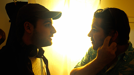 Two men face each other in front of a window where lots of natural light is entering a dimly lit room. Man on the left is wearing a hat and headphones around his neck. Man on right has his index finger up to his ear.