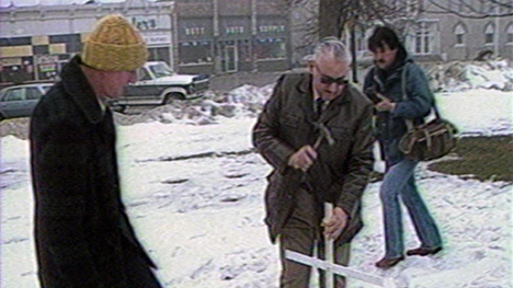 Three men stand in the snow in a town square. The oldest man in the middle is hammering a white cross into the ground while the others look on.
