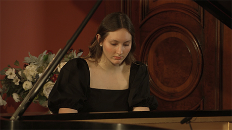 A young woman with shoulder length brown hair, wearing a black dress plays music on a grand piano.