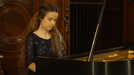 A young woman in sequin dress plays music on a grand piano.