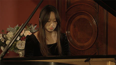 A young woman in a black dress plays music on a grand piano.