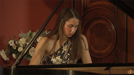 A young woman with long brown hair sits at a grand piano playing music.
