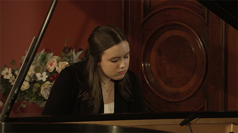 A young woman in a black and white outfit plays music on a grand piano.