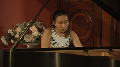 A young woman in an ivory floral dress plays music on a grand piano.