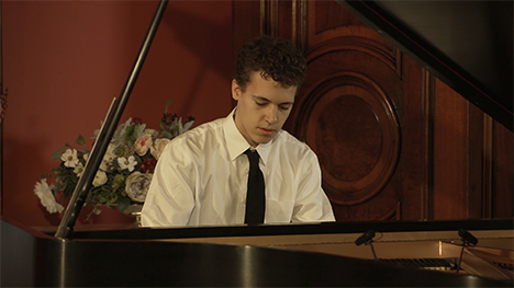 A young man in a white shirt and black tie sits playing at a grand piano.