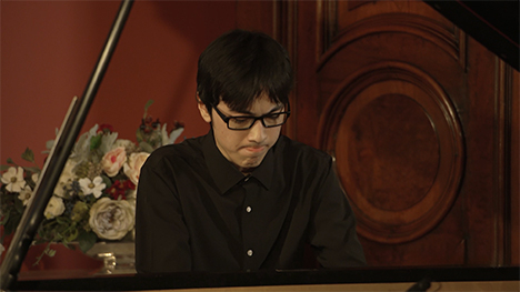 A young man in a black shirt and glasses sits playing at a grand piano.