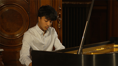 A young man in a white shirt sits playing at a grand piano.