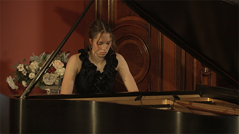 A young woman in a long black dress sits playing at a grand piano.