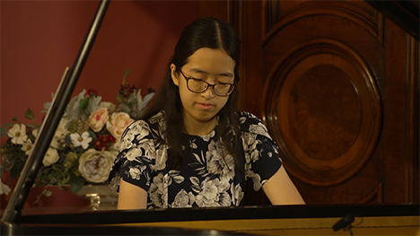 A young woman in a flowery patterned dress plays music on a grand piano.