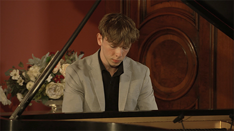 A young man in a black shirt and tan jacket sits playing at a grand piano.