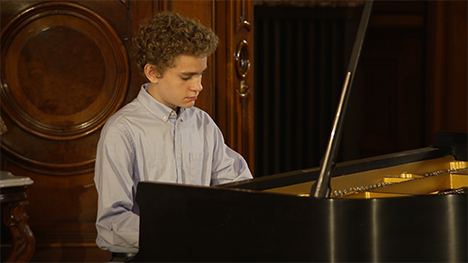 A young man in a light blue shirt sits playing at a grand piano.