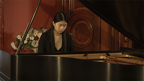 A young woman in a black dress plays music on a grand piano.
