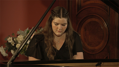 A young woman in a black dress plays music on a grand piano. 
