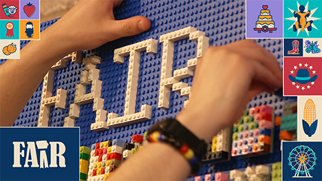 A person holds The LEGO Brick Wall Build with their left hand and adds another piece to the LEGO display with their right hand. White LEGO pieces spell out, "FAIR" against the blue LEGO base sheet. Quilt-like squares are displayed below.