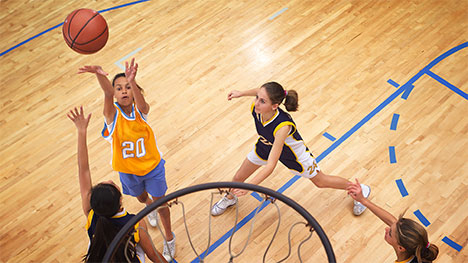 A girl basketball player shoots the basketball towards the hoop as players form the opposing team try to intercept.
