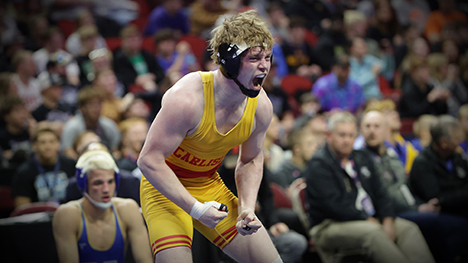A high school male wrestler in a yellow singlet belts out a yell and clenches his fists in front of wrestling spectators.