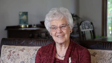 An older woman with white hair and glasses, sitting on a patterned sofa.