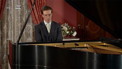 A young man in a suit sits playing at a grand piano.