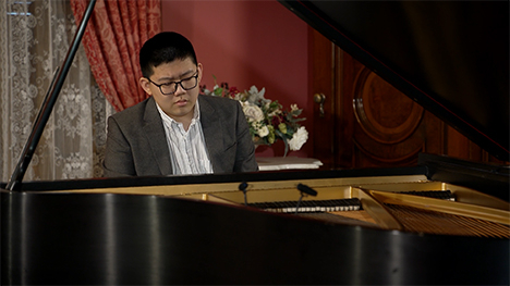 A young man in a suit sits playing at a grand piano.