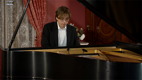 A young man in a suit sits playing at a grand piano.
