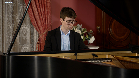 A young man in a suit sits playing at a grand piano.