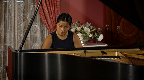 A young woman in a dress sits playing at a grand piano.