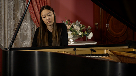 A young woman in a black dress sits playing at a grand piano.