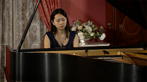 A young woman in a purple dress sits playing at a grand piano.