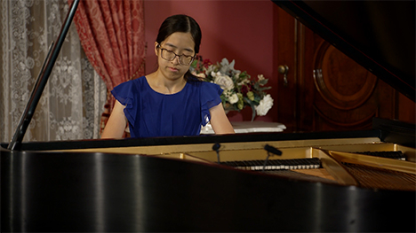 A young woman in a blue dress sits playing at a grand piano.