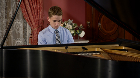 A young man in a shirt and tie sits playing at a grand piano.