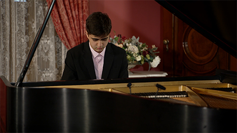 A young man in a suit sits playing at a grand piano.